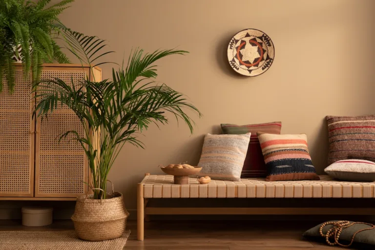 Shows a bench with pillows stage on top, large plants, and a privacy barrier all in front of a painted wall. The image is lit with a warm yellow light.