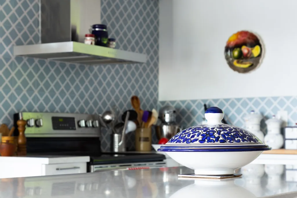 A blue container in front of a kitchen in the background with a colorful designed wall, also shows a kitchen hood, stove, and utensils.