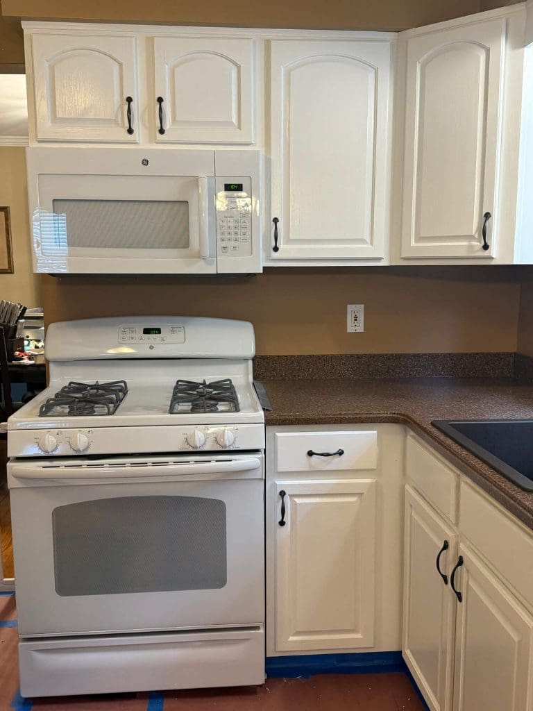 A kitchen with white cabinets, a white microwave, and a gas stove with a brown countertop and blue painter's tape on the floor.