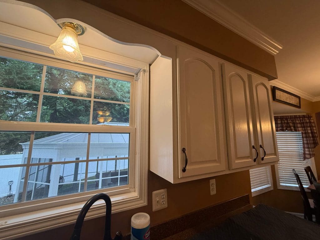 A kitchen with white cabinets, a window overlooking a screened porch, a decorative light fixture, and a countertop with a cleaning spray bottle and black faucet.