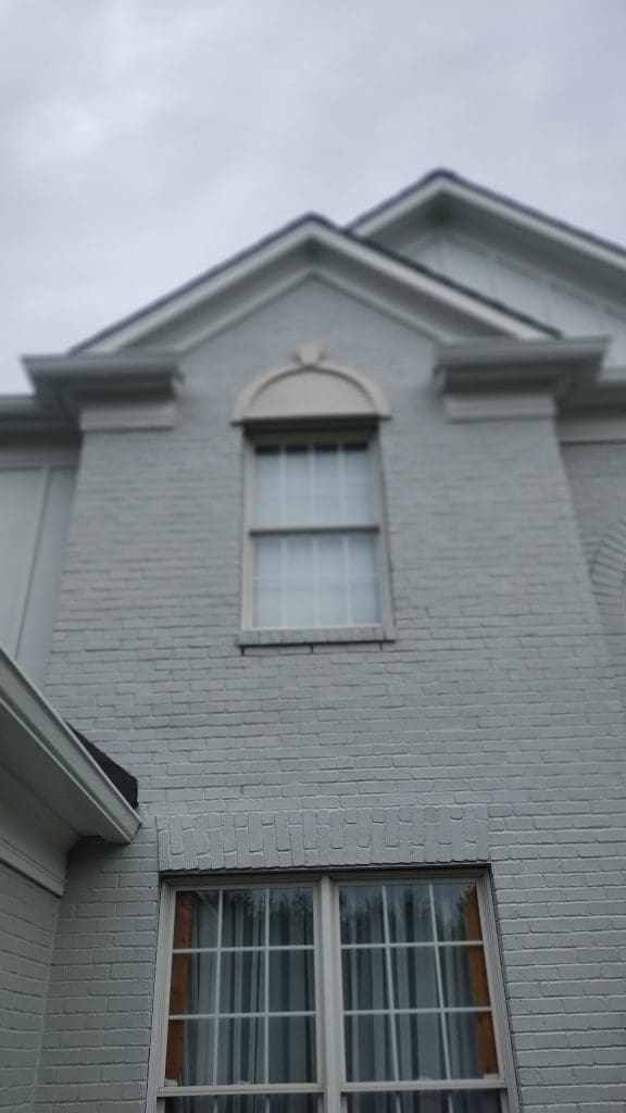 A gray brick house facade with two windows, one arched and one rectangular, under a cloudy sky.