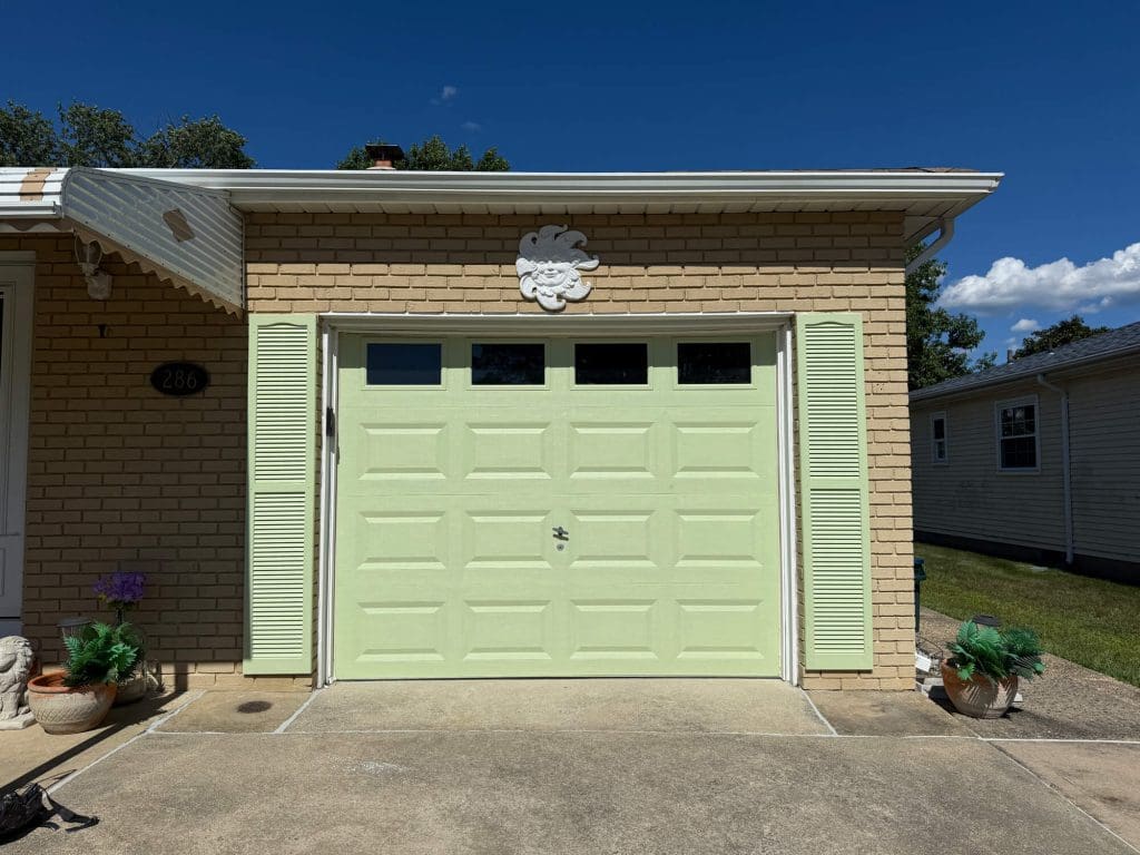 A beige brick house with a light green garage door, white shutters, a decorative sun ornament, and potted plants, under a clear blue sky.