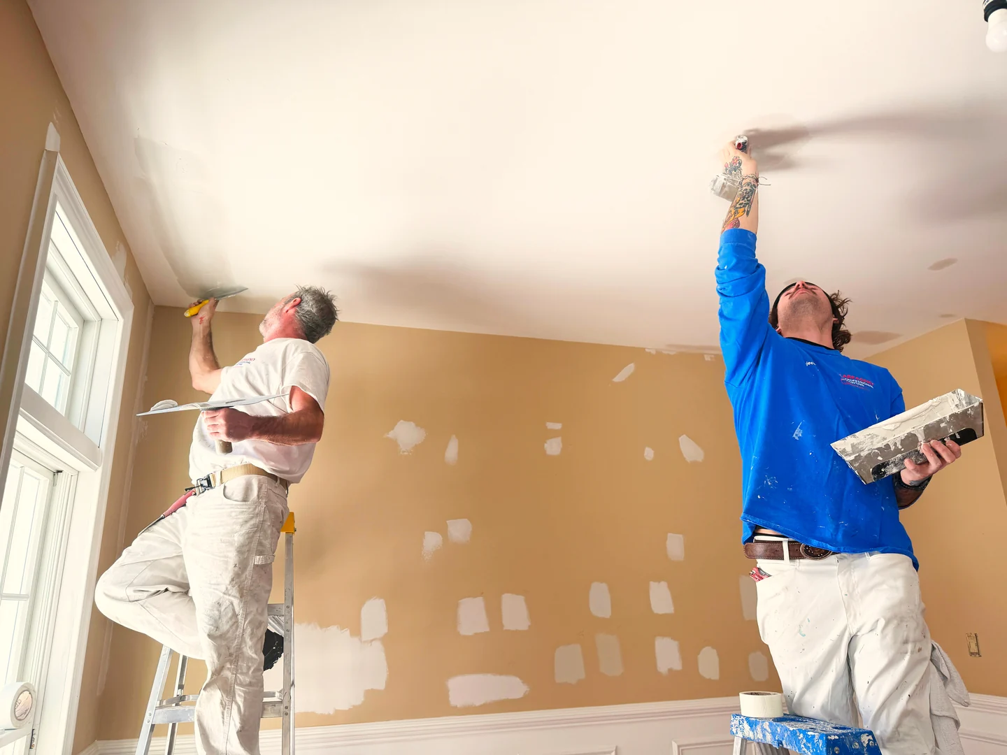 Two painters applying patching compound to a ceiling above walls with visible repairs.