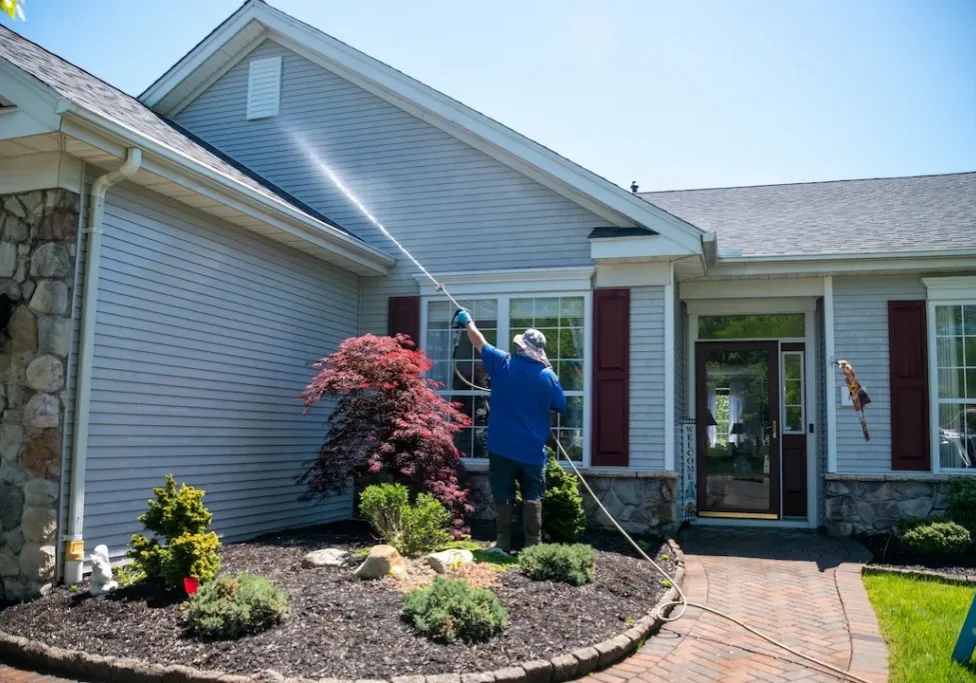 A caucasian man with a latex holding water sprayer wand power washing the siding of a house